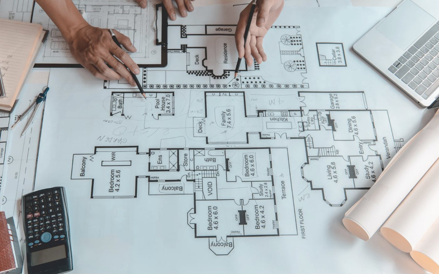 Two people marking up a house floor plan with pencils, beside rolled drawings